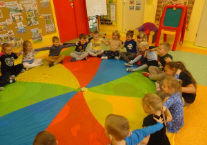 Grupa dzieci siedzi na brzegu chusty animacyjnej w siadzie skrzyżnym z dłońmi położonymi na ramionach kolegi bądź koleżanki siedzących po prawej i lewej stronie.